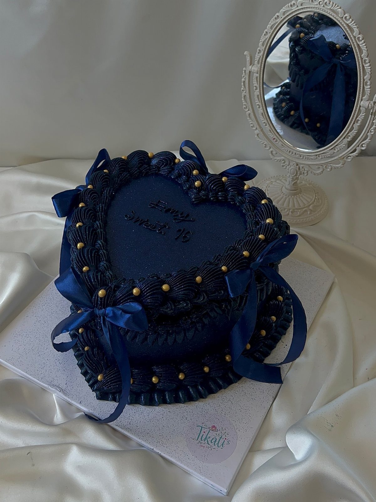 Heart-shaped dark blue cake with gold pearls and ribbons on white satin.