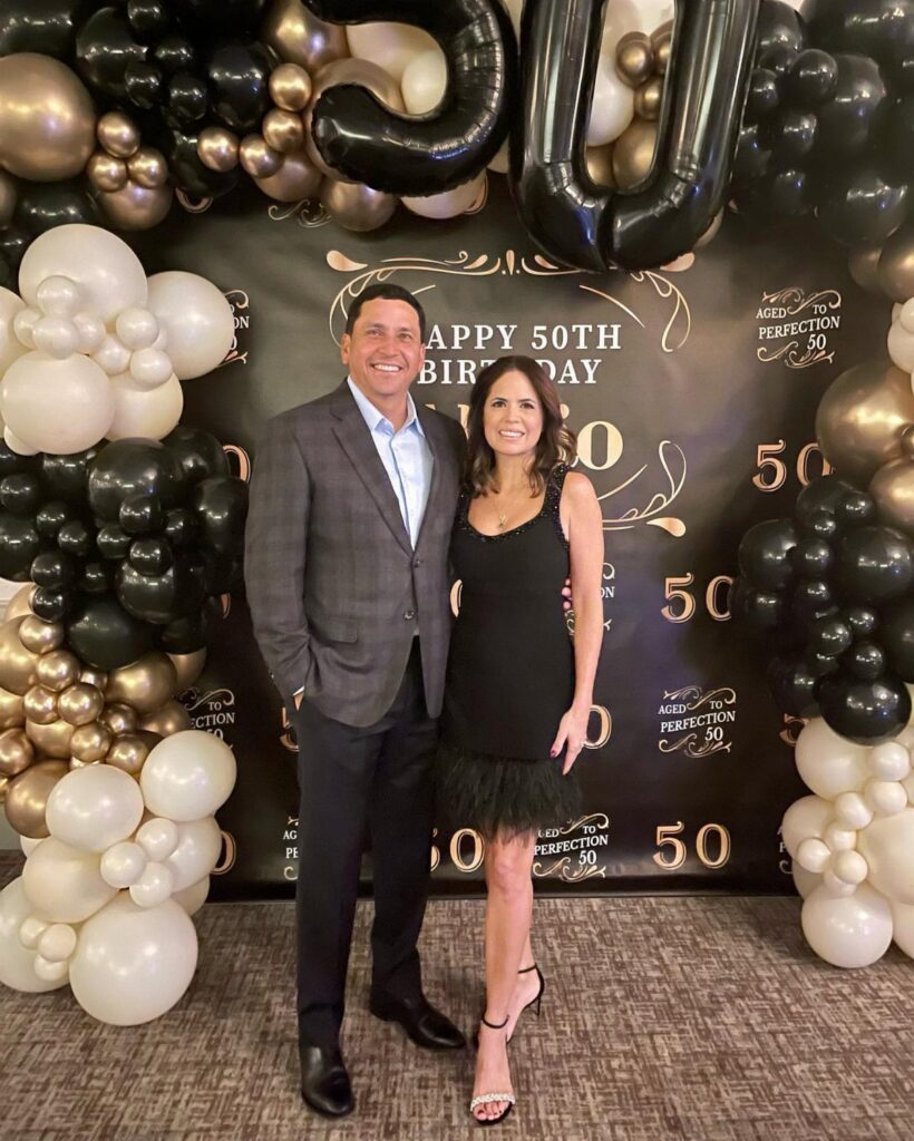Man and woman posing in formal attire before a 50th birthday balloon backdrop.