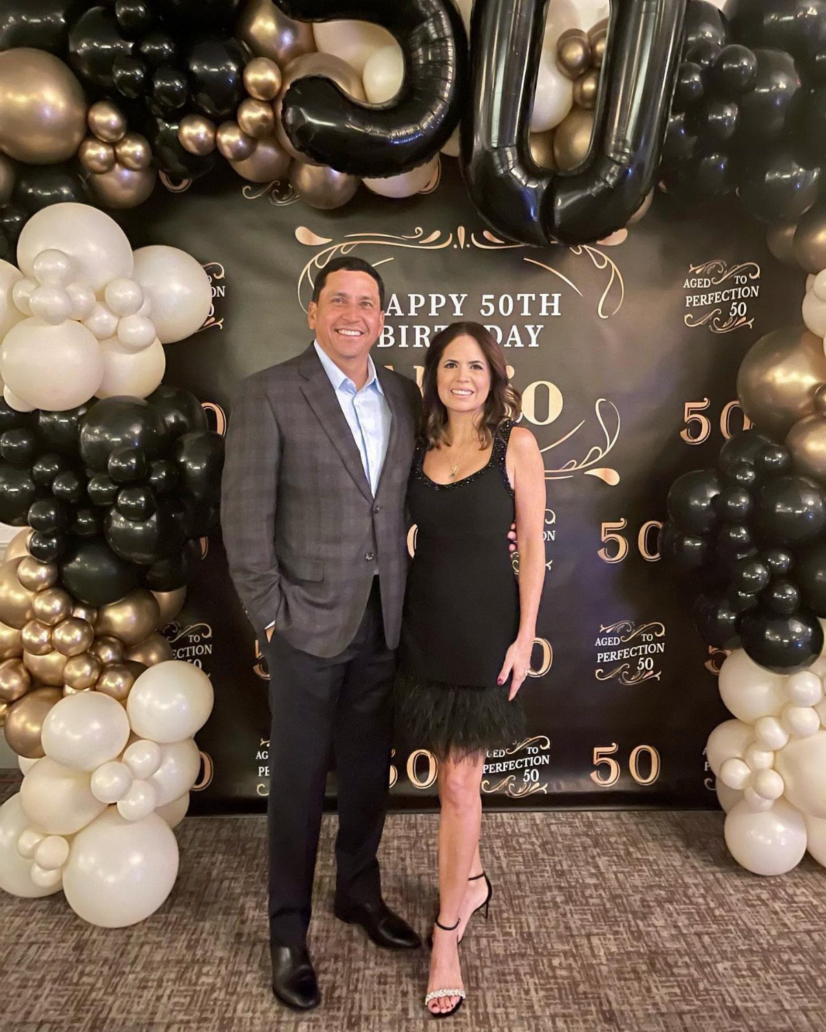Man and woman posing in formal attire before a 50th birthday balloon backdrop.