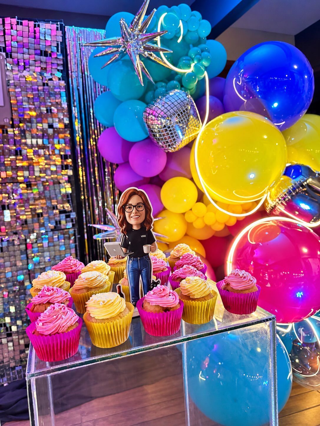 Colorful cupcakes on a clear stand before a vibrant balloon arch and sequin wall.