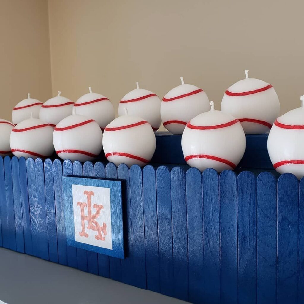 Baseball-themed white candles with red stitching arranged on a blue picket fence.