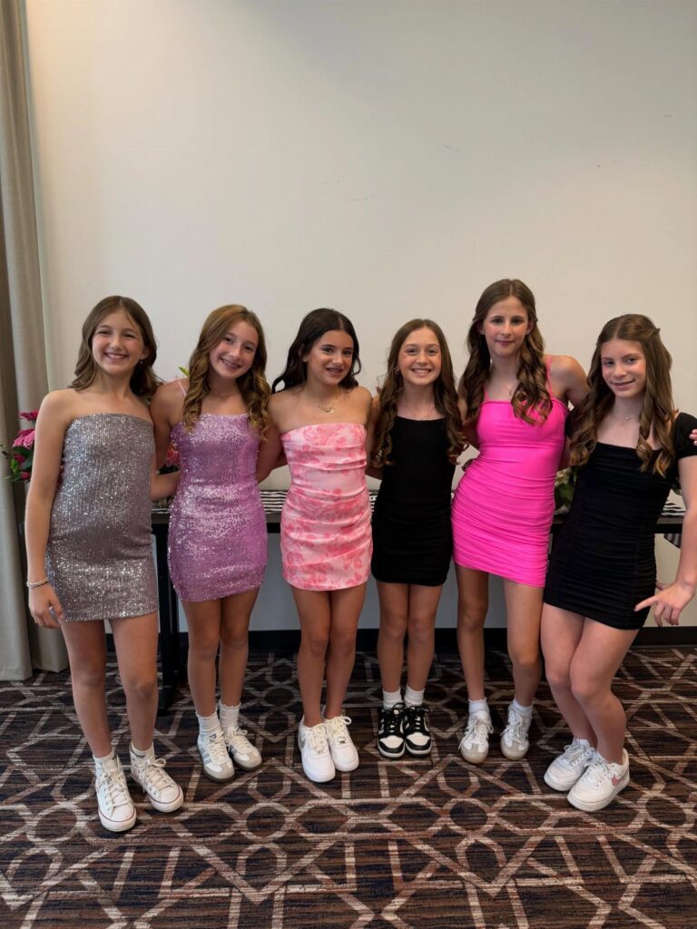 Six girls in colorful sequined party dresses and white sneakers posing together indoors.