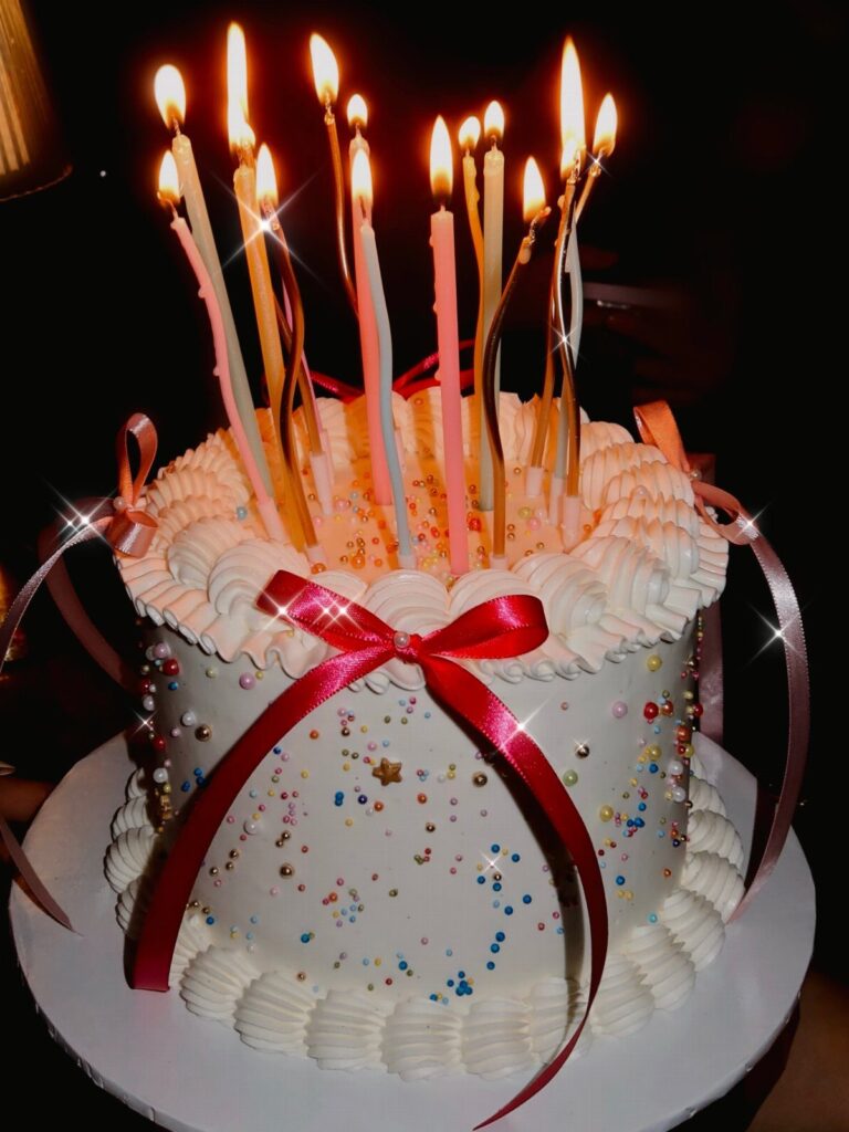 White frosted birthday cake with a red bow and many lit colorful candles.