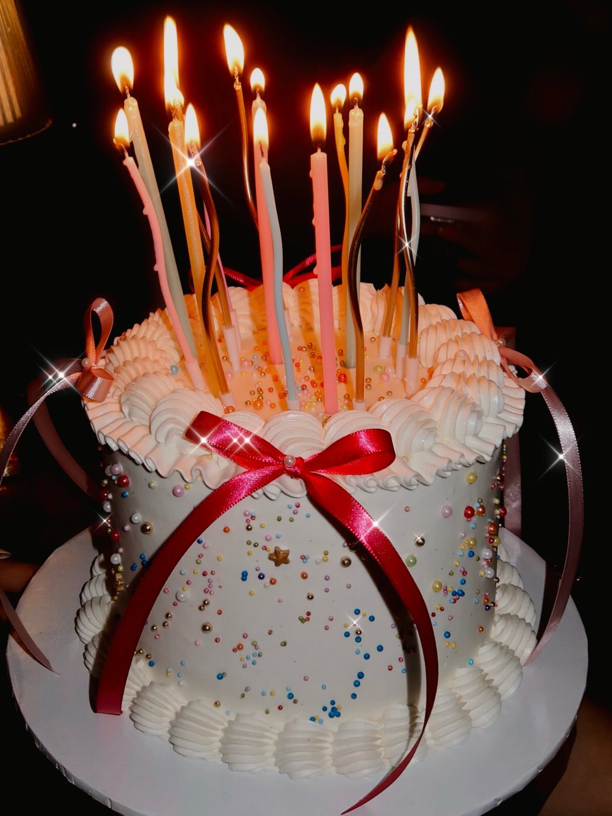 White frosted birthday cake with a red bow and many lit colorful candles.