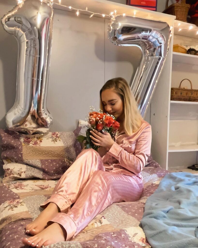 Young woman in pink satin pajamas holding red roses next to 17 balloons