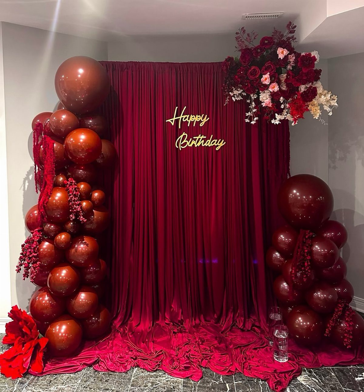 Red velvet birthday backdrop with metallic red balloon arches and floral arrangements.
