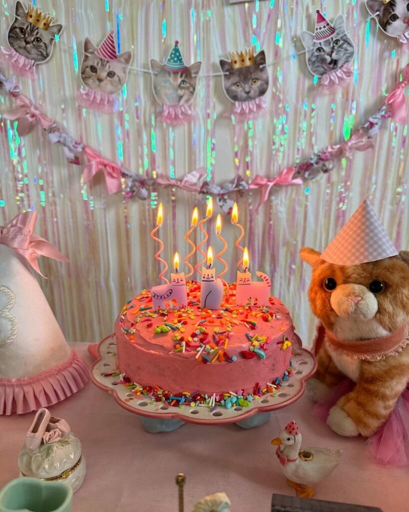 Pink birthday cake with sprinkles and a cat plushie wearing a party hat.