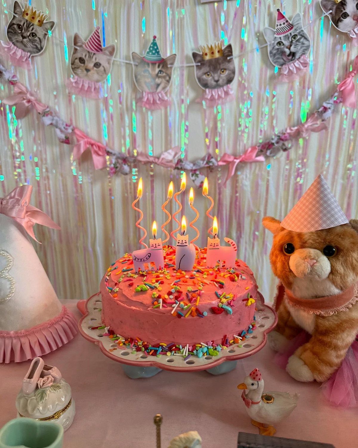 Pink birthday cake with sprinkles and a cat plushie wearing a party hat.