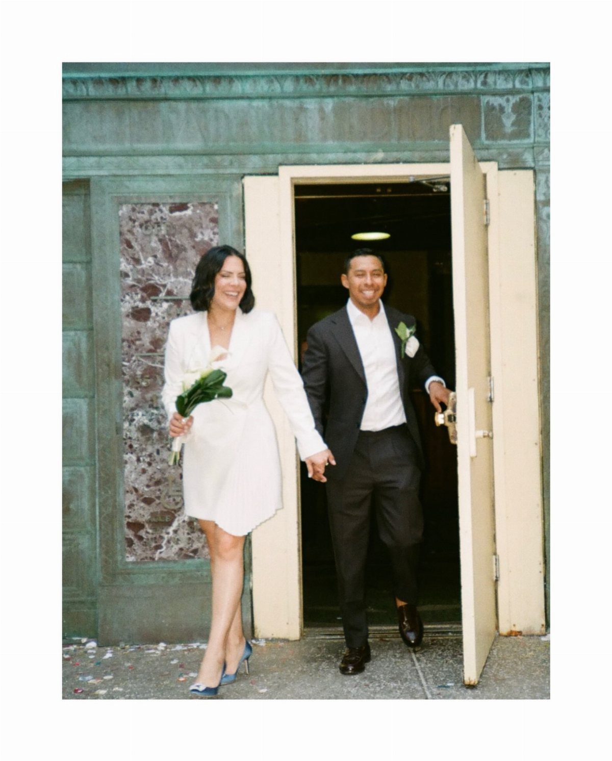 Newlywed couple in a white blazer dress and black suit exiting a building.