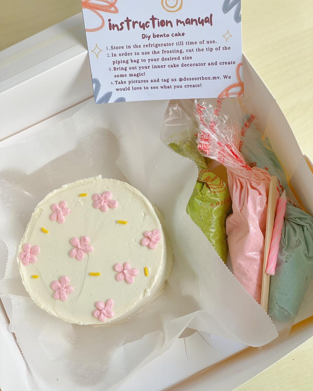 Small white cake with pink flowers and colorful frosting piping bags in a box.