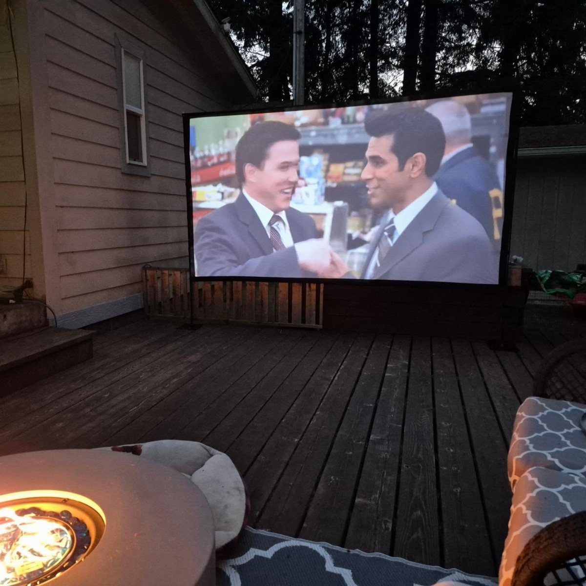 Projector screen showing a movie on a wooden deck with a fire pit.
