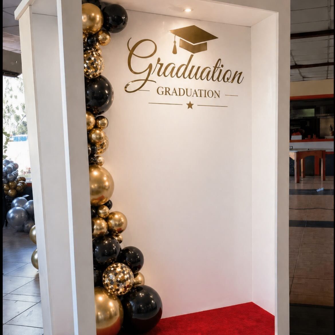 Graduation photo booth backdrop with gold and black balloons and red carpet.