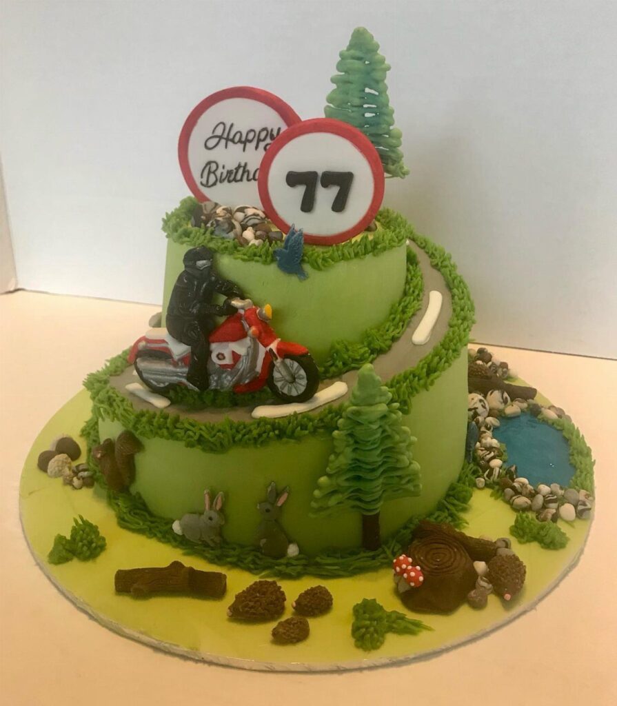 Two-tiered green hill cake with a motorcyclist, trees, and birthday signs.