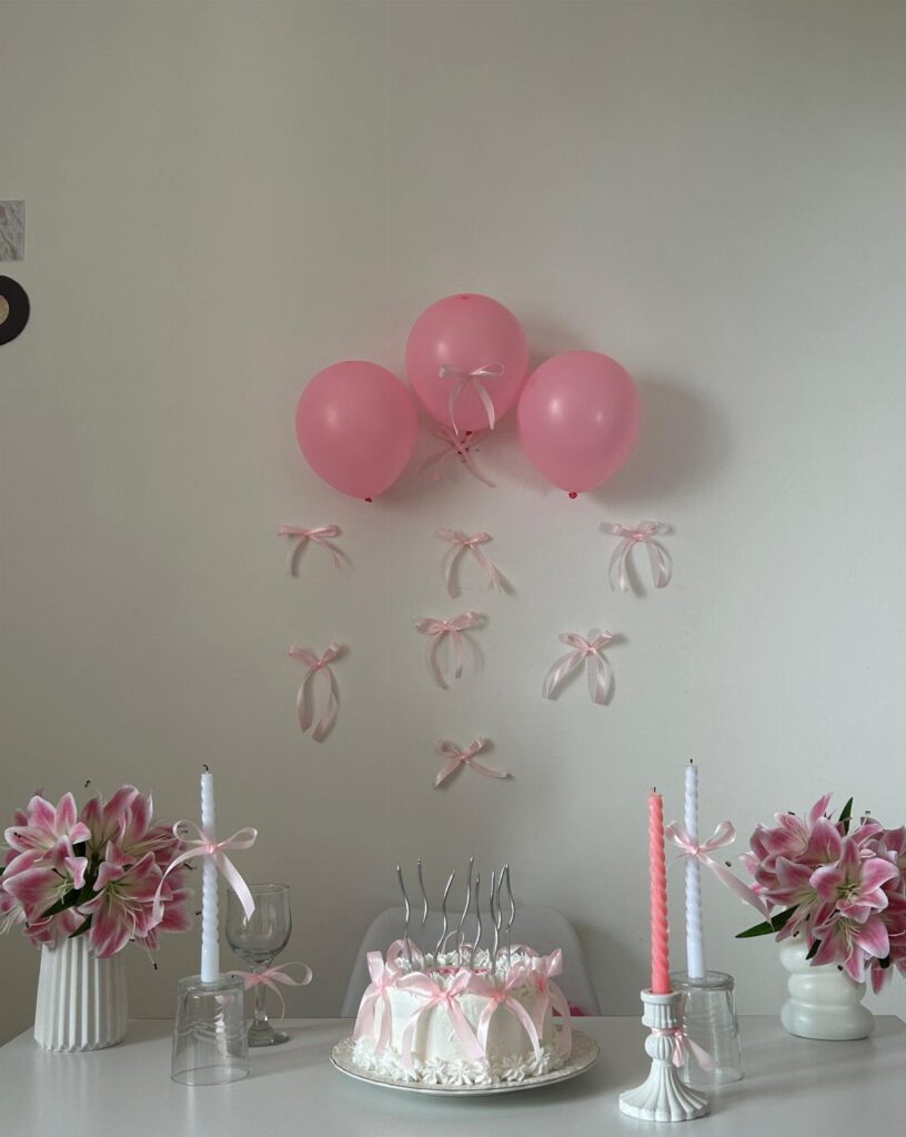 Pink birthday party setup with cake, balloons, lilies, and candles on a white table
