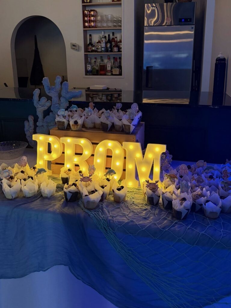 Glowing PROM letters and blue coral decorations on a table with cupcakes and netting.