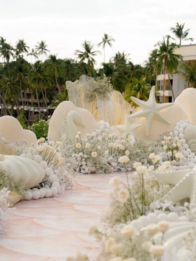 Wedding aisle decorated with giant white seashells, starfish, and white flowers on a beach.